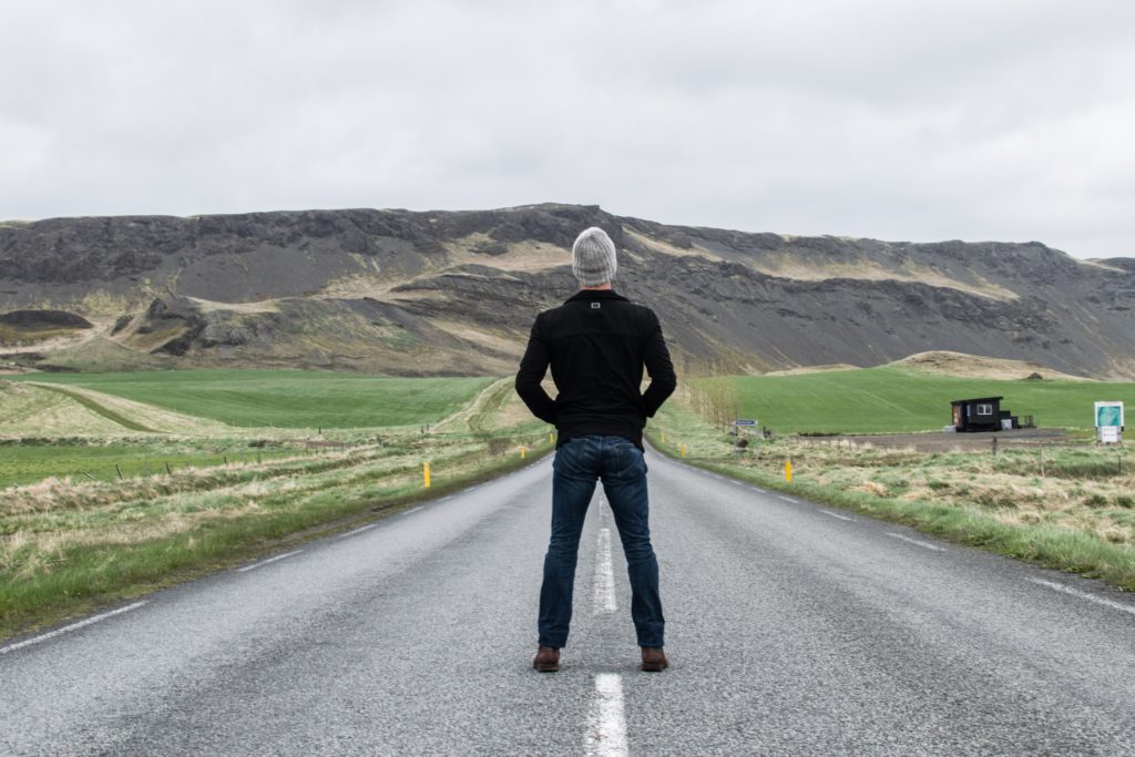 A man is standing on an empty road with countryside around and hills ahead. He is lost.