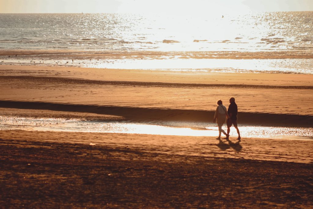 2 people walking along a beach, on orange coloured sad, at sunset