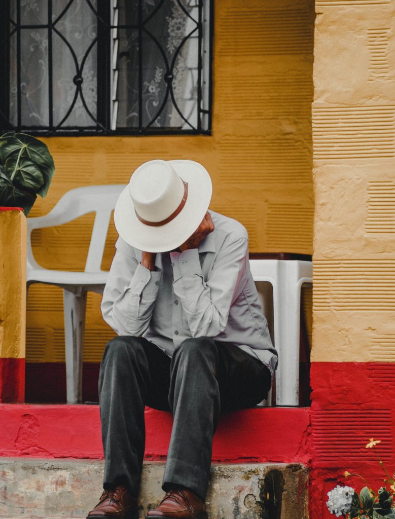 Smartly dressed man sitting at the front of his house on a veranda, with his head in his hands, and a wide brimmed hat on.